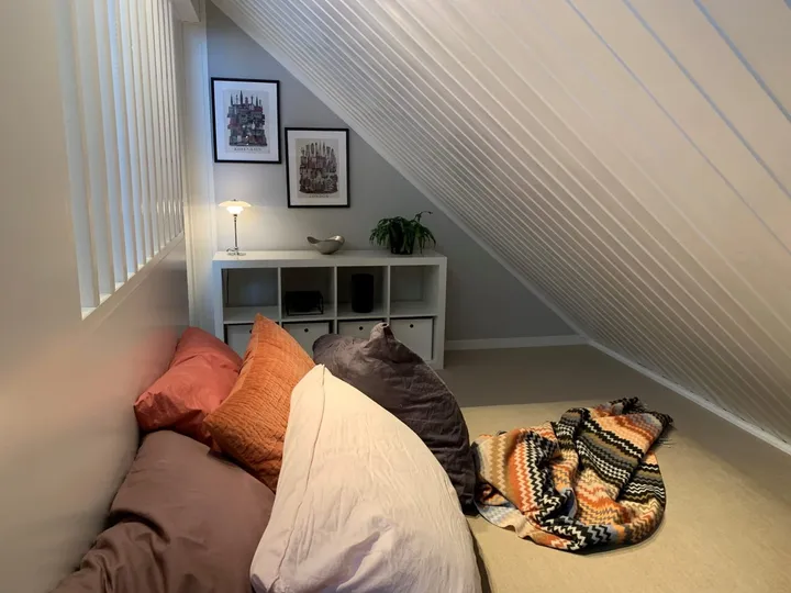 Loftstue: loftsrom med hvitt skråtak, puter og pledd i oransje og brunt