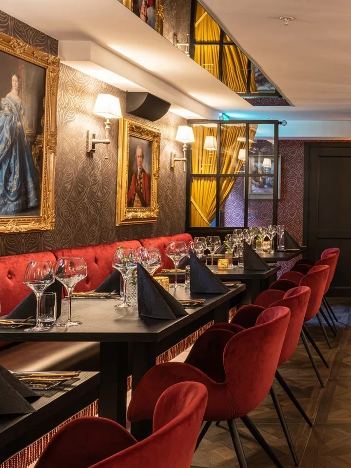 Restaurant Alma – Tønsberg: dekket langbord med røde stoler og benk, med portretter i gullrammer på mønstret vegg