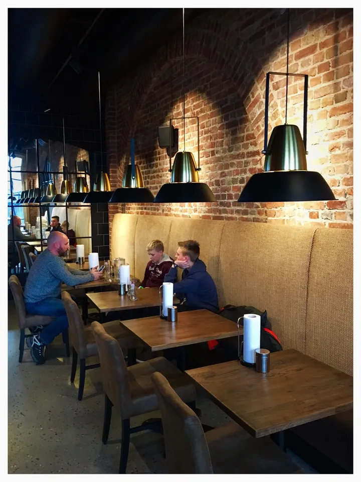 Sørsida Grill & BBQ – Hønefoss: gjester ved trebord foran en murvegg, med beige benk og svarte pendellamper