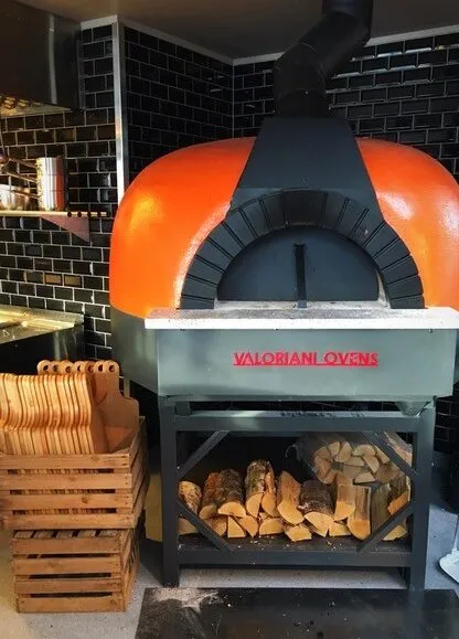 Tavernan Alta: oransje pizzaovn med påskriften «Valoriani Ovens» og en vedstabel under
