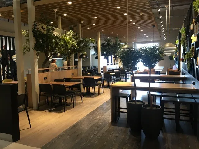 Coop Cafe Rudshøgda: åpen spisesal med trebord, trær i store krukker og tretak med lamper