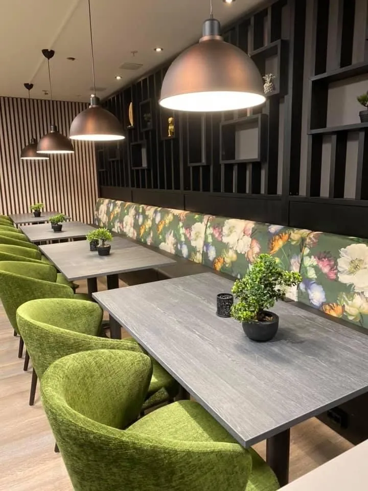 Coop Cafe Steinkjer: grønne polstrede stoler ved grå bord og en benk med blomstermønster foran en mørk lamellvegg
