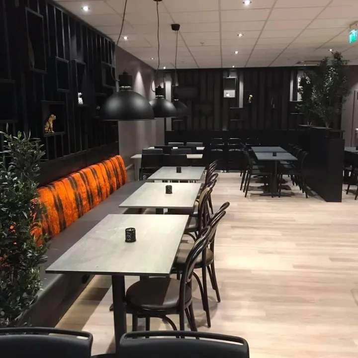 Coop Cafe Steinkjer: lang rekke grå bord og sorte stoler i en mørk spisesal, med oransje benk langs veggen