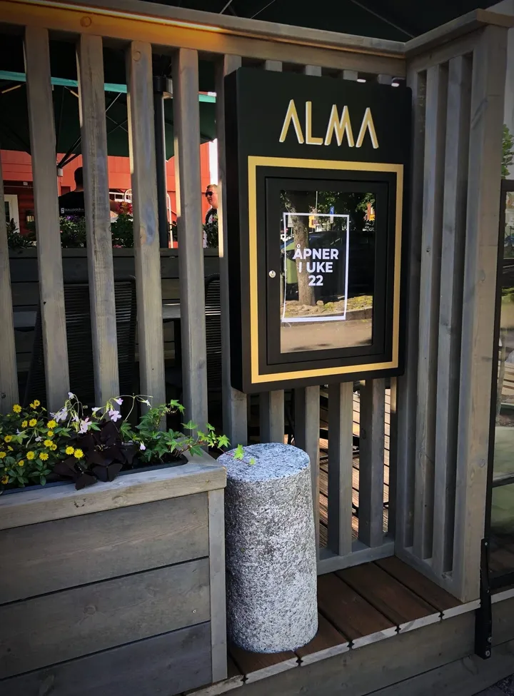 Restaurant Alma – Tønsberg: gul plakat i ramme med teksten «Åpner uke 22» på et trigjerde ved en blomsterkasse