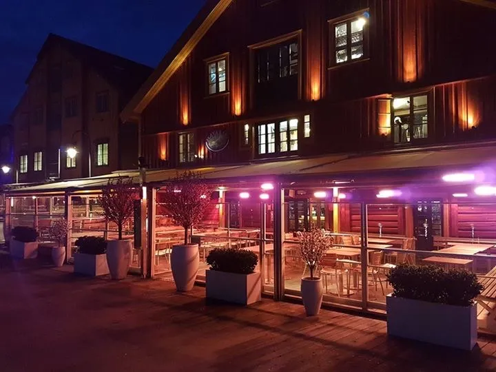 Havariet: rødbrun trebygning med opplyst uteservering og planter i kasser på kvelden