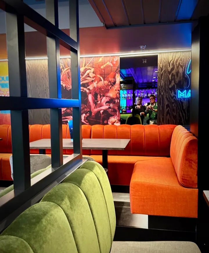 Lille Havariet: oransje sofa og grønne fløyelspuffer bak et sort rutenett, med neonlys i bakgrunnen