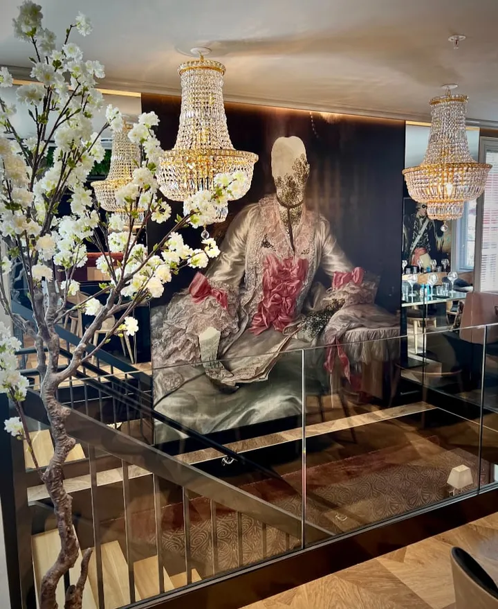 Alma restaurant: veggmotiv av en kledd figur med lysekroner og blomstrende grener foran et glassrekkverk