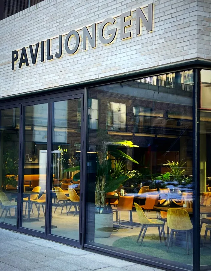 Paviljongen Drammen: glassfasade med svarte rammer mot en uteplass med planter og gule stoler