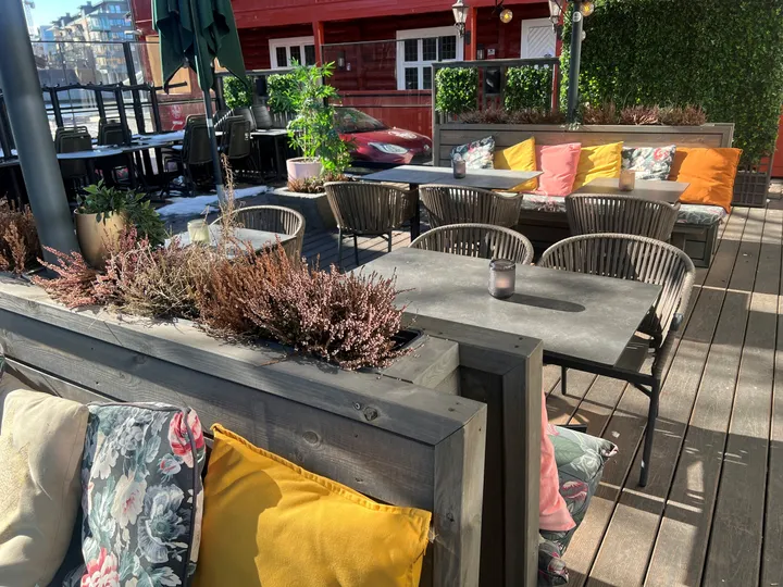 Restaurant Alma – Tønsberg: uteservering på et tredekk med grå trebenker, flettede stoler, fargerike puter og lyng i plantekasser