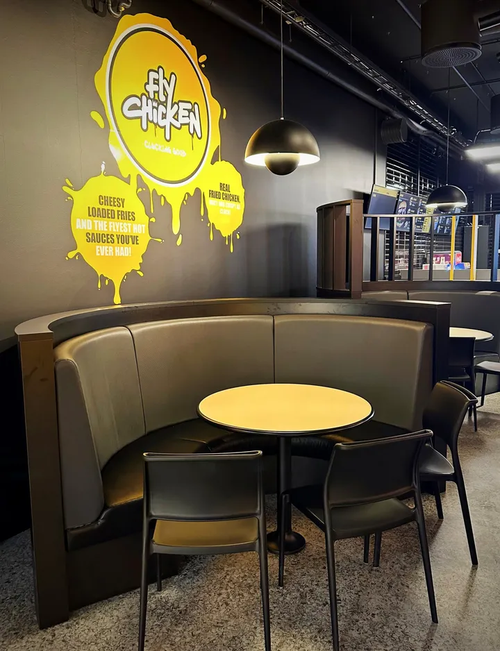 Fly Chicken: halvrund sofa i grått rundt et gult bord, under en svart lampe og en gul logo med teksten «Fly Chicken» på veggen
