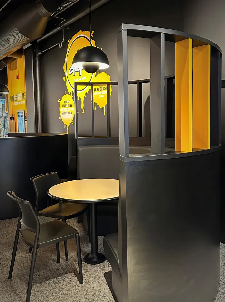 Fly Chicken: sittebås med gule detaljer og runde gule bord, med gule plakater på den mørke veggen