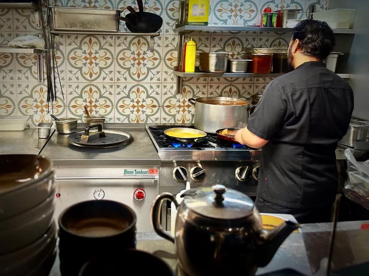 India Gate – Bergen: kokk ved gassbluss og tandoori-ovn i et kjøkken med mønstrede fliser i grønt og oker